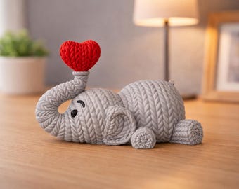May include: A gray elephant figurine with a textured knit-like surface pattern. The elephant is lying down with its trunk curled upward, holding a bright red heart that also features a textured knit-like appearance. It has black eyes.