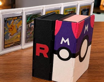 May include: A black deck box featuring a red letter R and a purple, white, and pink deck box with a Master Ball design and the letter M. Both boxes are rectangular with a textured finish, standing upright on a wooden surface with card displays.