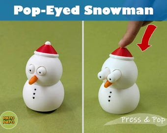 May include: A white snowman figurine with a red hat, orange carrot nose, and three black buttons. The toy features a mechanism where pressing the top of the hat causes the two white eyes to pop outward. The text Pop-Eyed Snowman and Press & Pop appear.