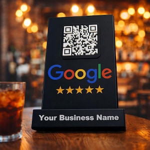 Könnte beinhalten: Schwarzes Tischschild mit einem QR-Code, dem Google-Logo und fünf goldenen Sternen. Das Schild zeigt auch den Text "Your Business Name" in Weiß. Der Hintergrund ist verschwommen mit warmer Beleuchtung.