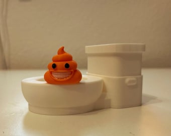 May include: A small orange figurine shaped like a coiled pile with black eyes and a wide white toothy grin, resting on the rim of a white plastic toilet model. The toilet features a tank, a bowl, and a flush handle on the side.