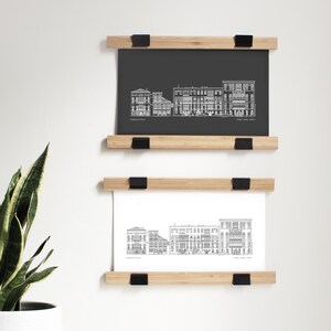 Può includere: Due stampe architettoniche incorniciate. La stampa superiore è su sfondo grigio scuro, quella inferiore è bianca. Entrambe le stampe presentano un disegno a tratto bianco di una facciata di edificio. Le cornici sono in legno chiaro con clip nere.