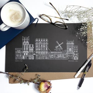 May include: A black and white line drawing of Parisian buildings, including the Moulin Rouge, on black paper. The artwork is surrounded by a cup of coffee, glasses, pens, and dried flowers. The text "Moulin Rouge, Paris" is visible.