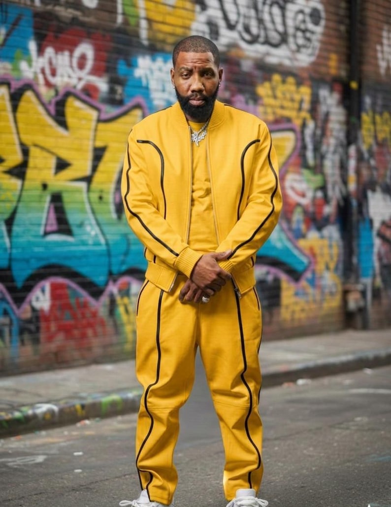May include: A yellow tracksuit with black trim is worn by a person standing on a city street. The jacket has a zipper and the pants have a drawstring. The background features colorful graffiti art.