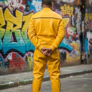 May include: A person wearing a bright yellow leather jacket and matching pants with black trim, standing in front of a graffiti-covered wall. The jacket has a horizontal black stripe across the upper back.