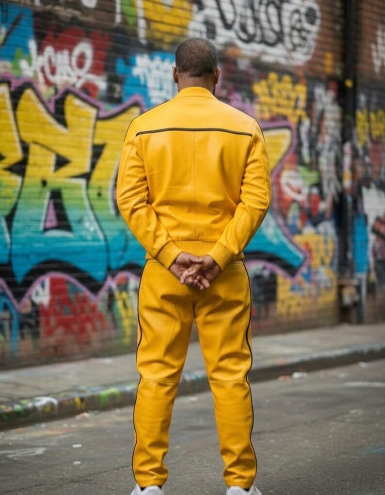 May include: A person wearing a bright yellow leather jacket and matching pants with black trim, standing in front of a graffiti-covered wall. The jacket has a horizontal black stripe across the upper back.