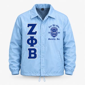 May include: A light blue coach jacket featuring a snap-button front closure, a collar, and long sleeves with elastic cuffs. The front displays large dark blue Greek letters ZΦB vertically on the left and a circular Zeta Phi Beta 1920 logo on the right.
