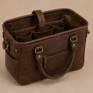May include: A brown leather tote bag with multiple internal pockets and a zipped front pocket. The bag has a top handle and a shoulder strap attachment. The hardware is brass-coloured, and the bag appears to be well-made.