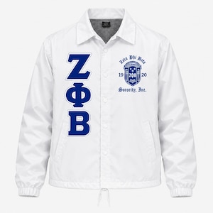 May include: A white long-sleeve coach jacket with a collar and snap-button front. The left side features the blue Greek letters ZΦB vertically. The right side displays a blue crest with the text Zeta Phi Beta 1920 Sorority, Inc.