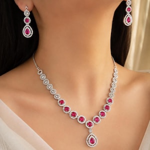 May include: A silver necklace and earring set. The necklace features a series of red, oval gemstones encircled by clear, round stones. The earrings mirror the design, with three red gemstones and clear stones. The jewellery is designed to be worn together.