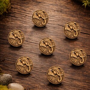May include: Seven antique brass-colored cabinet knobs with an elephant design. Each knob features a detailed relief of an elephant's head within a circular frame. The knobs are arranged on a rustic wooden surface, showcasing their intricate details.