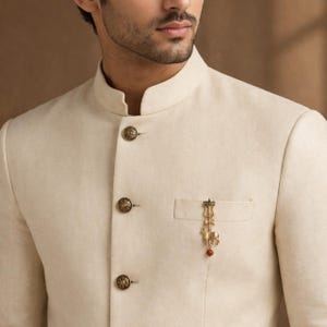 May include: Ivory-colored jacket with ornate, gold-toned buttons and a small pocket. A decorative gold brooch with a red bead and green accents adorns the pocket. The jacket has a mandarin collar.