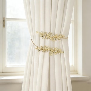 May include: Two gold-colored curtain tiebacks with a leaf and pearl design. The tiebacks are wrapped around white curtains, adding a decorative touch to the window treatment. The leaf design is detailed with small, sparkling accents.