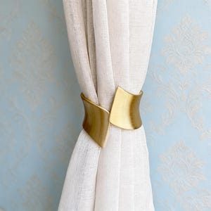 May include: A gold-coloured metal curtain tieback. The tieback is wrapped around a sheer white curtain. The tieback has a curved design with a brushed metal finish. The background is a light blue wall with a white floral pattern.