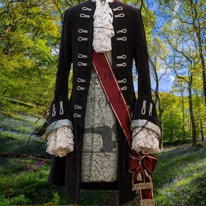 May include: A black velvet coat with silver buttons, paired with a patterned vest, a white ruffled cravat, and a burgundy sash. The sleeves feature lace cuffs, and a matching bow with tassels completes the ensemble. This historical costume evokes a sense of regal elegance.