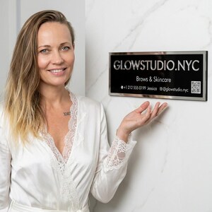 Op de afbeelding: Een vrouw in een witte badjas staat naast een zwart en zilveren bord met de tekst GLOWSTUDIO.NYC, met daaronder de woorden Brows & Skincare. Het bord bevat ook een telefoonnummer, de naam Jessica en een e-mailadres.