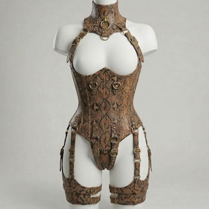 Peut inclure: Un body marron à motif peau de serpent avec un col montant, des bretelles et des jarretelles. Le vêtement comporte des anneaux et des boucles en métal doré. Le body est présenté sur un mannequin blanc.