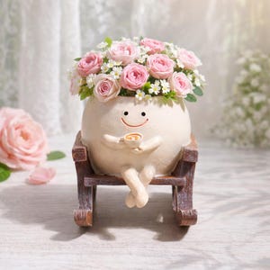 Puede incluir: Una maceta caprichosa, de color crema, con forma de personaje sonriente, sosteniendo una taza y sentado en una mecedora de madera. La maceta está llena de rosas rosas y margaritas blancas. Un artículo decorativo encantador.