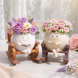 Op de afbeelding: Twee grillige bloempotten in de vorm van lachende personages, een in een houten schommelstoel, de andere in een houten stoel. De ene houdt een boek vast, de andere een kopje. Beide zijn gevuld met kleurrijke bloemen, waaronder rozen en lavendel.