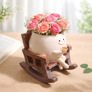 May include: A cream-colored, round, smiling character planter sitting in a brown wooden rocking chair, holding a small cup. The planter is filled with a bouquet of pink roses and white baby's breath flowers, creating a decorative floral arrangement.