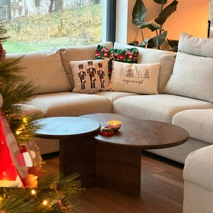 Op de afbeelding: Een gezellige woonkamer met een lichtgekleurde hoekbank versierd met decoratieve kussens. Twee donkere, ronde salontafels van hout staan voor de bank. Een kerstboom met ornamenten staat op de voorgrond.