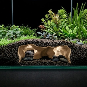 Puede incluir: Una estructura de cueva para acuario de color beige en forma de túnel, incrustada en un sustrato granular de color marrón oscuro. La cueva presenta múltiples aberturas y cámaras interiores, rodeada de plantas acuáticas verdes y piedras.