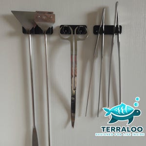 Könnte beinhalten: Set von Aquarienwerkzeugen aus Edelstahl, einschließlich Schaber, Spatel, Schere und Pinzette, an einer weißen Wand montiert. Die Werkzeuge werden von schwarzen Haltern gehalten. Das Terraloo-Logo befindet sich unten rechts.