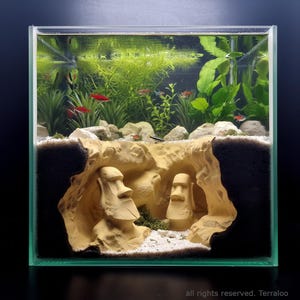 Peut inclure: Un aquarium carré en verre présentant une vue en coupe d'une grotte sablonneuse contenant deux statues Moaï beiges. La partie supérieure comprend des plantes aquatiques vertes, du gravier blanc et plusieurs petits poissons rouges et argentés.