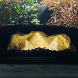 Könnte beinhalten: Ein Diorama mit drei hellgelben Pyramidenstrukturen, die aus dem Inneren einer dunklen, strukturierten höhlenartigen Umhüllung beleuchtet werden. Die Pyramiden sind von dunklem, körnigem Material umgeben, das an Erde erinnert.