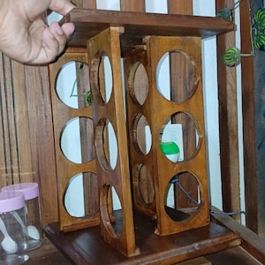 Può includere: Un portabottiglie in legno marrone scuro. Presenta diverse aperture circolari per contenere i barattoli di spezie. Il design comprende un ripiano superiore e una base. Una mano tiene il ripiano superiore. Il portabottiglie è su una superficie di legno.