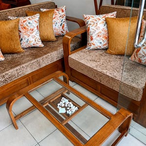 Puede incluir: Juego de sofás de madera con cojines estampados y una mesa de centro con tapa de cristal. Los cojines del sofá son de color marrón texturizado, y los cojines presentan diseños de hojas naranjas y blancas. La mesa de centro tiene un estante inferior con piedras decorativas.