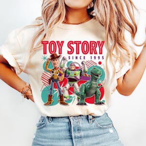 Peut inclure: Un t-shirt couleur cr&egrave;me avec une impression graphique de Woody, Buzz l'&Eacute;clair et Rex de Toy Story. Le texte indique TOY STORY SINCE 1995 en rouge et noir. Le motif comprend des cercles bleu sarcelle, des lignes diagonales rouges et des formes.