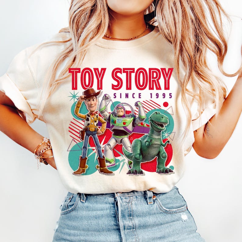 Peut inclure: Un t-shirt couleur cr&egrave;me avec une impression graphique de Woody, Buzz l'&Eacute;clair et Rex de Toy Story. Le texte indique TOY STORY SINCE 1995 en rouge et noir. Le motif comprend des cercles bleu sarcelle, des lignes diagonales rouges et des formes.