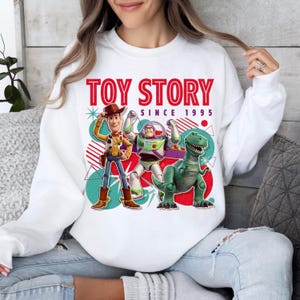 Peut inclure: Un sweat-shirt blanc &agrave; col rond avec une impression graphique de Woody, Buzz l'&Eacute;clair et Rex de Toy Story. Le texte indique TOY STORY SINCE 1995 en rouge et noir. Le motif comprend des formes g&eacute;om&eacute;triques color&eacute;es, des cercles et des &eacute;toiles.