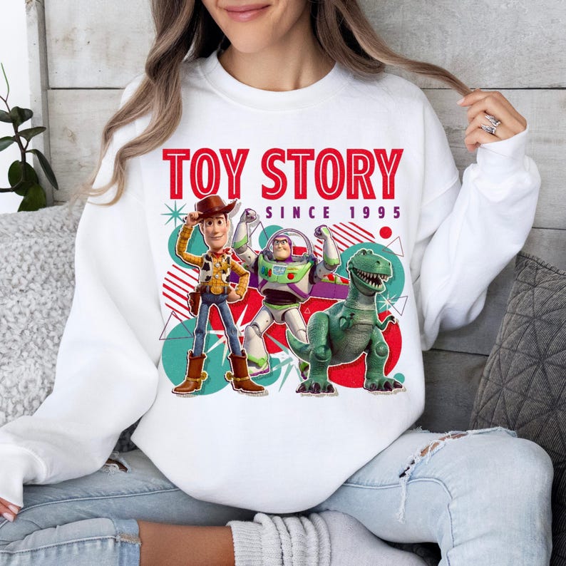 Peut inclure: Un sweat-shirt blanc &agrave; col rond avec une impression graphique de Woody, Buzz l'&Eacute;clair et Rex de Toy Story. Le texte indique TOY STORY SINCE 1995 en rouge et noir. Le motif comprend des formes g&eacute;om&eacute;triques color&eacute;es, des cercles et des &eacute;toiles.