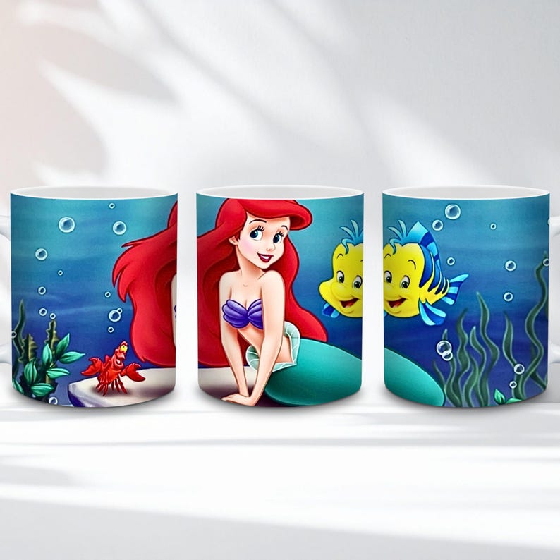Op de afbeelding: Witte keramische mok met Ariel de zeemeermin met rood haar, een paarse top en een zeegroene staart, samen met Botje en Sebastiaan. De mok heeft een blauwe onderwaterachtergrond met bellen en zeewier.