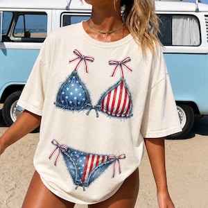 以下が含まれることがあります： オフホワイトの半袖Tシャツで、デニム風のビキニのグラフィックプリントが施されています。ビキニの上下には、青い背景に白い星、赤と白のストライプが描かれており、赤と白のリボンがアクセントになっています。