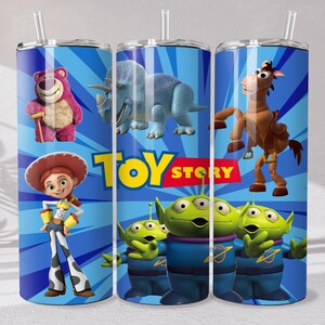 May include: A cylindrical tumbler with a straw featuring a blue background with radiating lines and the Toy Story logo. Characters include a pink bear, a blue triceratops, a brown horse, a cowgirl, and three green three-eyed aliens in blue suits.