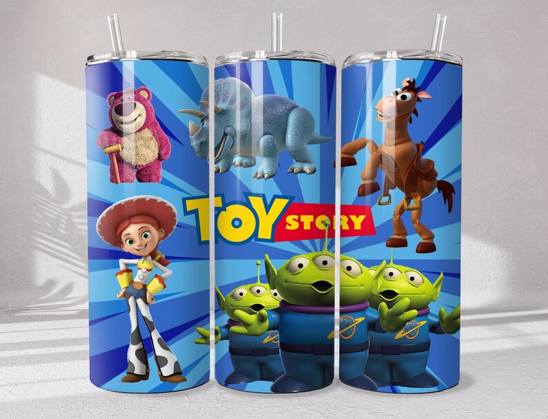 May include: A cylindrical tumbler with a straw featuring a blue background with radiating lines and the Toy Story logo. Characters include a pink bear, a blue triceratops, a brown horse, a cowgirl, and three green three-eyed aliens in blue suits.