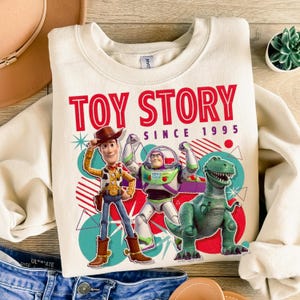 Peut inclure: Un sweat-shirt &agrave; col rond couleur cr&egrave;me avec une impression graphique de Woody, Buzz l'&Eacute;clair et Rex de Toy Story. Le texte indique TOY STORY SINCE 1995 en rouge et violet. Le motif comprend des formes g&eacute;om&eacute;triques et des lignes color&eacute;es.