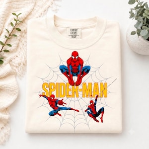 May include: A cream-colored t-shirt featuring a graphic print of three Spider-Man figures in red and blue suits posed against a light gray spiderweb background. The text SPIDER-MAN is printed in bold yellow letters across the center of the design.