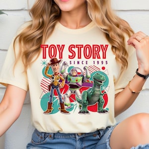 Peut inclure: Un t-shirt couleur cr&egrave;me avec une impression graphique de Woody, Buzz l'&Eacute;clair et Rex de Toy Story. Le texte indique TOY STORY SINCE 1995 en lettres rouges. Le motif comprend des formes g&eacute;om&eacute;triques, des cercles et des &eacute;toiles bleu sarcelle et rouges.