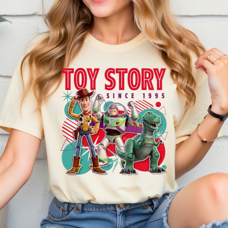Peut inclure: Un t-shirt couleur cr&egrave;me avec une impression graphique de Woody, Buzz l'&Eacute;clair et Rex de Toy Story. Le texte indique TOY STORY SINCE 1995 en lettres rouges. Le motif comprend des formes g&eacute;om&eacute;triques, des cercles et des &eacute;toiles bleu sarcelle et rouges.