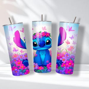 May include: Three cylindrical tumblers featuring a blue alien character with large ears and a flower crown. The design includes a vibrant garden of pink, purple, and blue flowers with scattered butterflies against a light blue sky background.