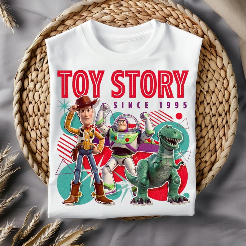 Peut inclure: Un t-shirt blanc avec une impression graphique de Woody, Buzz l'&Eacute;clair et Rex de Toy Story. Le texte indique TOY STORY SINCE 1995 en lettres rouges et noires. Le motif comprend des formes g&eacute;om&eacute;triques color&eacute;es, des cercles et des lignes en arri&egrave;re-plan.
