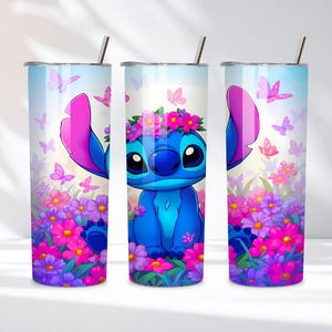May include: Three cylindrical tumblers with metal straws featuring a blue alien character with large ears wearing a pink flower crown. The design includes a vibrant field of pink, purple, and magenta flowers with scattered pink butterflies against a light blue sky.