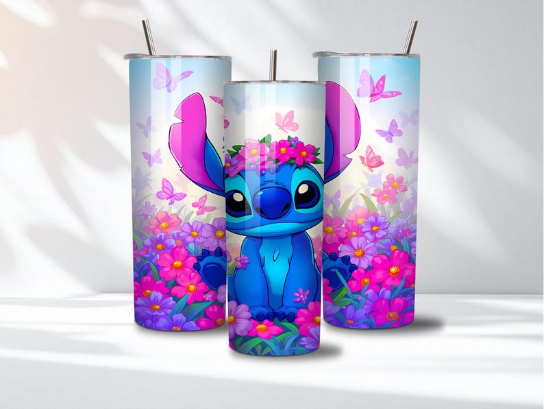 May include: Three cylindrical stainless steel tumblers with metal straws, featuring a blue alien character wearing a pink flower crown, surrounded by vibrant pink and purple flowers and butterflies against a light blue and white gradient background.