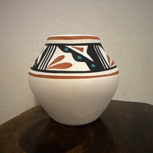 May include: A white ceramic vase with a rounded body and a wide opening. The rim is decorated with a black, white, and orange geometric pattern. The vase has a traditional Southwestern design.