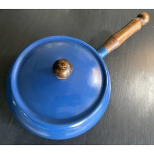 May include: A blue saucepan with a matching lid and a wooden handle. The lid has a wooden knob. The handle is attached to the pan with a blue metal piece. The pan is sitting on a dark surface.