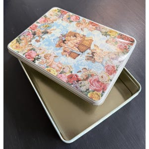 May include: A rectangular metal tin with a lid featuring a romantic design. The lid showcases cherubs, roses, and other flowers against a light blue background. The open tin reveals a gold interior.